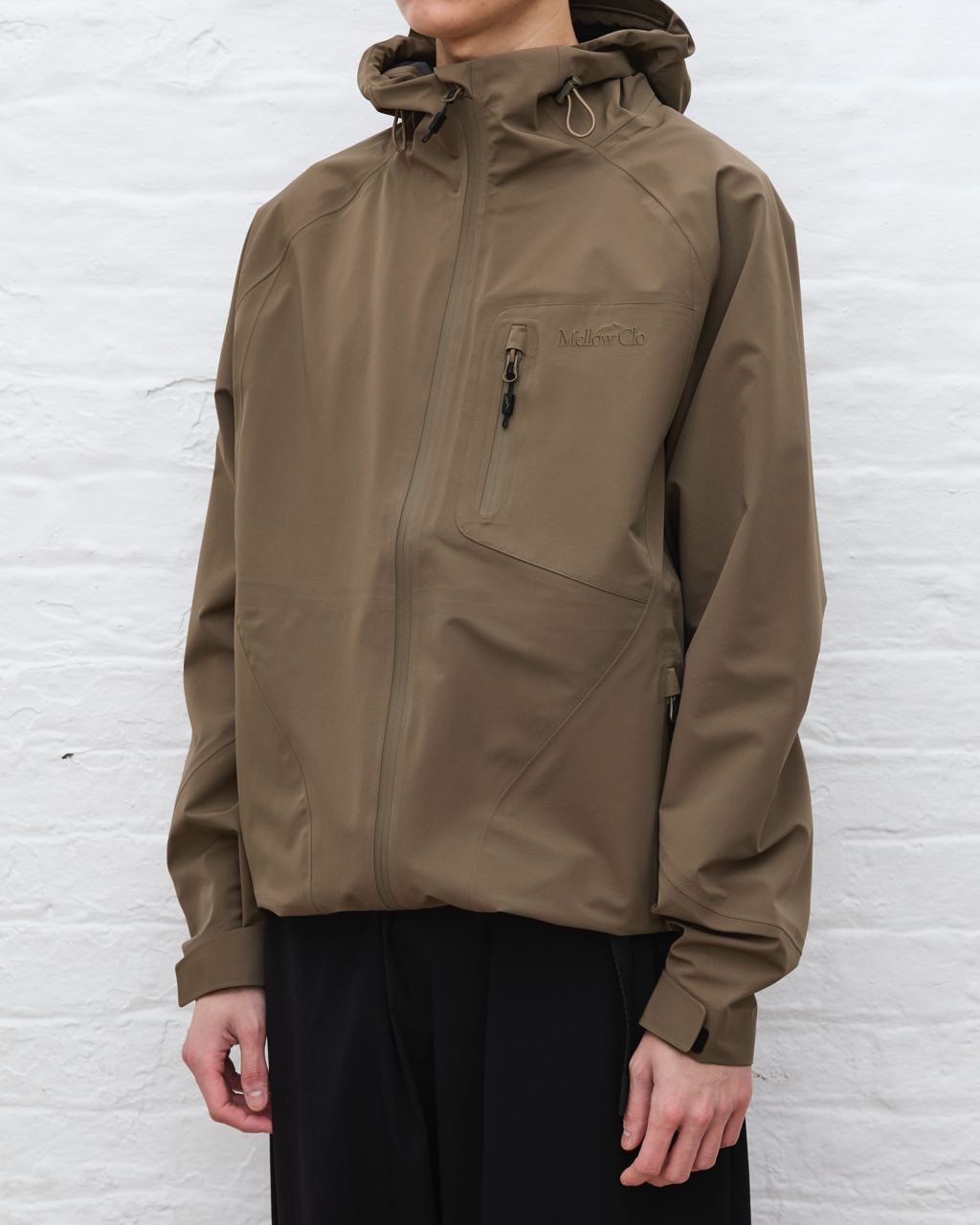 Person wearing a brown jacket with a brand logo against a white brick wall.