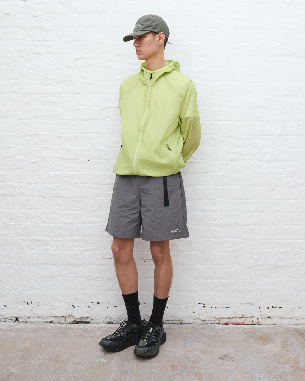 Person wearing a light green jacket, gray shorts, and black shoes against a white brick wall.