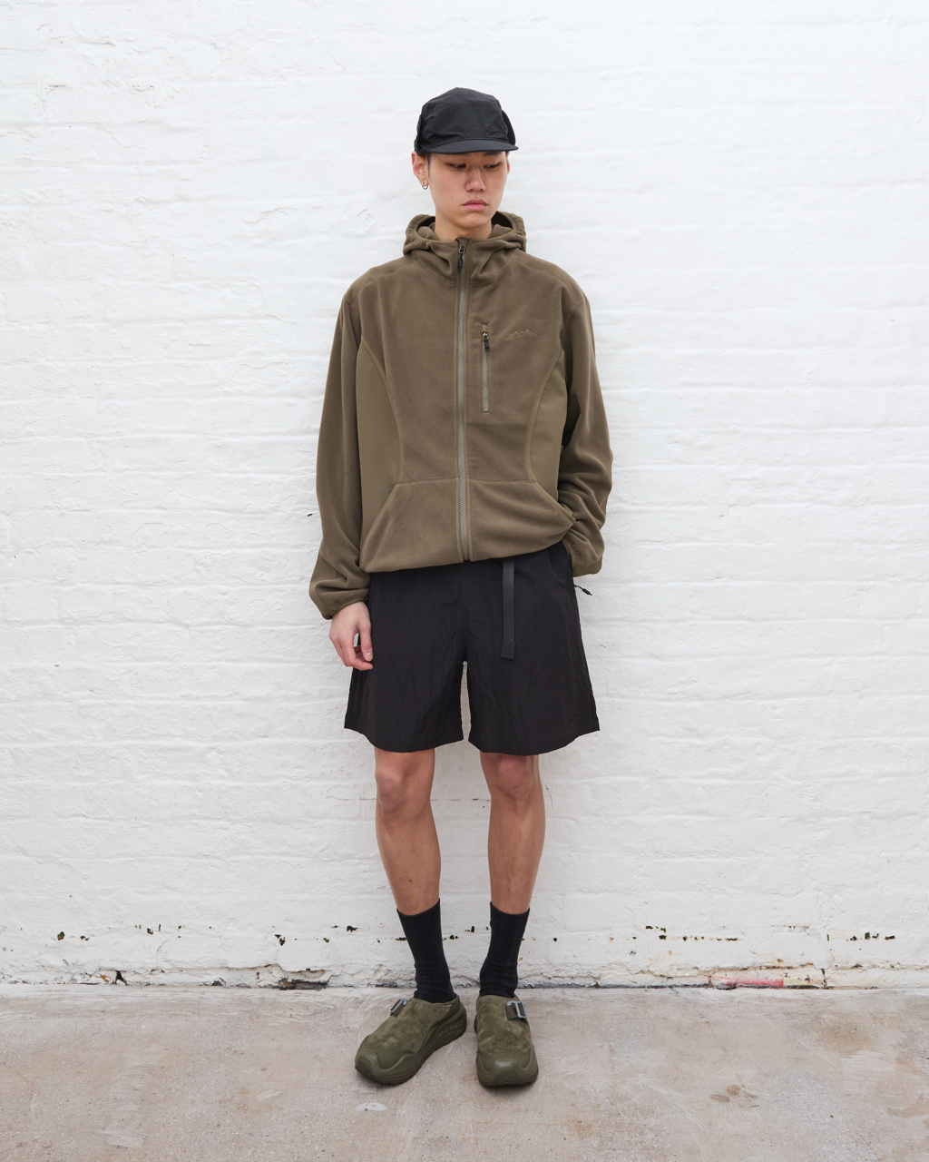 Person wearing a brown jacket, black shorts, and green shoes against a white brick wall.