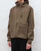Person wearing a brown jacket with a brand logo against a white brick wall.