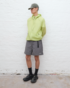 Person wearing a light green jacket, gray shorts, and black shoes against a white brick wall.